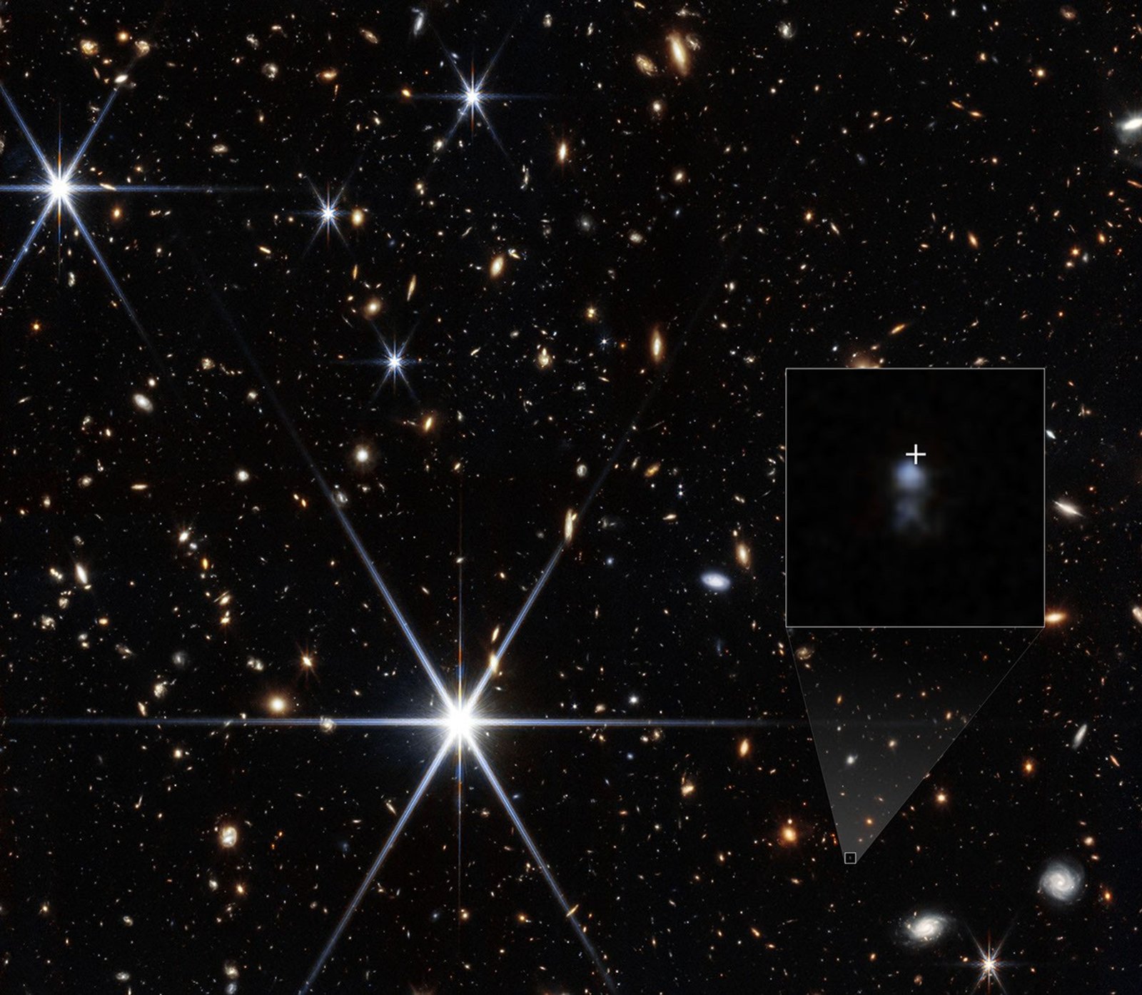 A field full of small galaxies of all shapes against the blackness of space. A few bright white stars show 8 prominent diffraction spikes. A small galaxy at lower right is highlighted with a white box. A shaded triangle extends upward to the corners of a larger box showing a zoom in of the region. Two fuzzy, light blue blobs are at center, with a larger and brighter one at top. Just above the top blob is a white crosshair showing the location where a fast radio burst was detected.