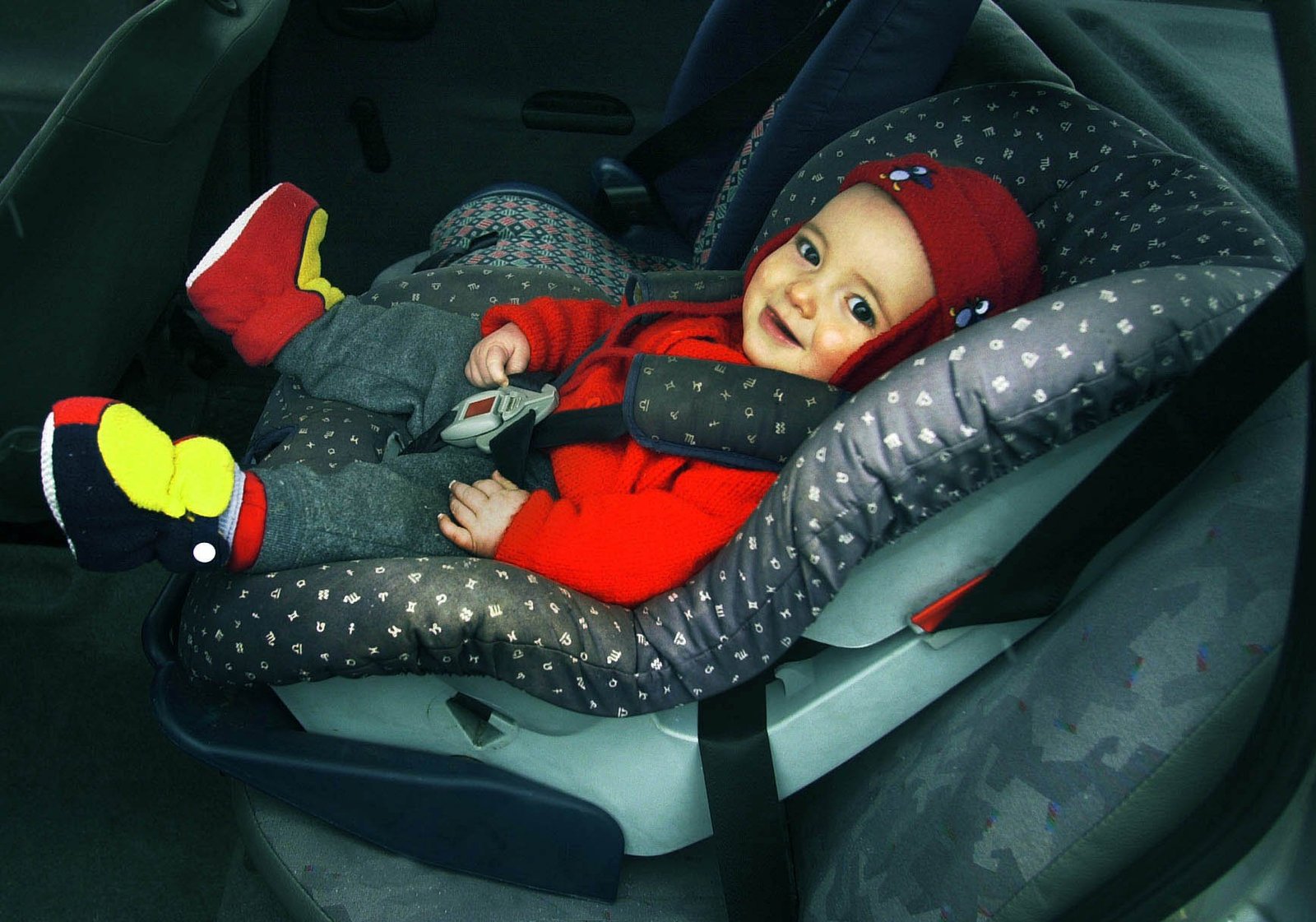 Baby in red hat and red and yellow shoes looks at camera from car seat.