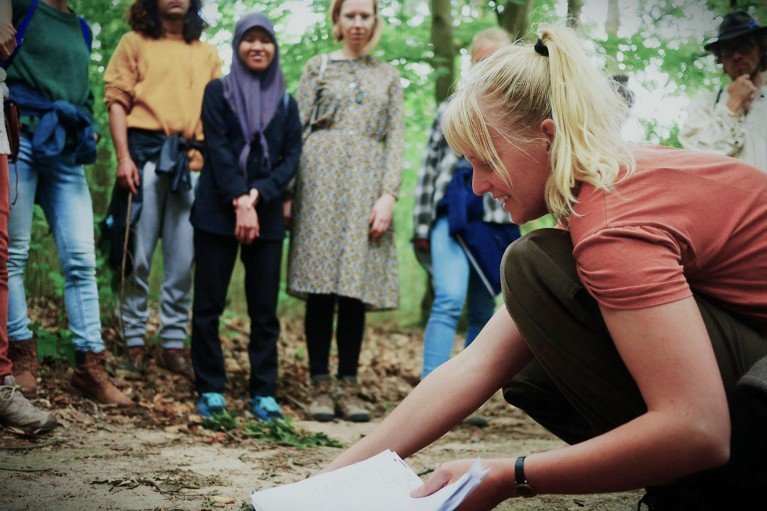 Lian Kasper crouches down in the foreground while holding paperwork and smiling as she talks to a group of students standing around her in a forest in Wageningen in the Netherlands.