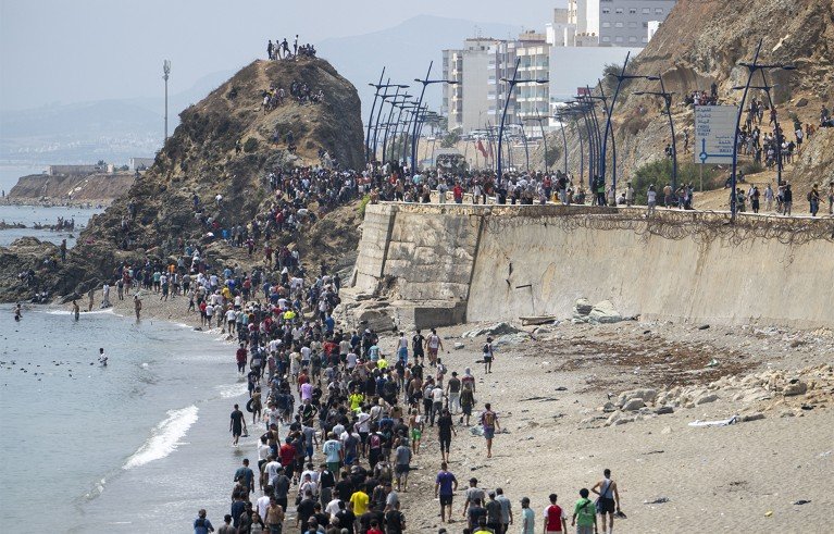 Migrants crowding a beach and coastal border area at Ceuta, Spain. Headline reports more than 48,300 migrants returned to Morocco, with fences, rocky cliffs, dense crowds.
