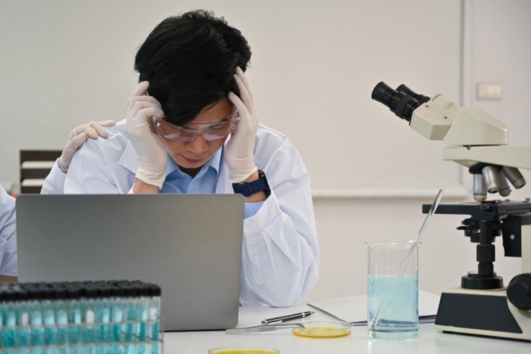 A laboratory worker in protective gear and a lab coat sits at a laptop beside a microscope and test tubes with his head in his hands