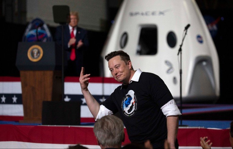 Elon Musk speaking at a SpaceX event, with a spacecraft capsule behind him and Donald Trump at a podium bearing the presidential seal.