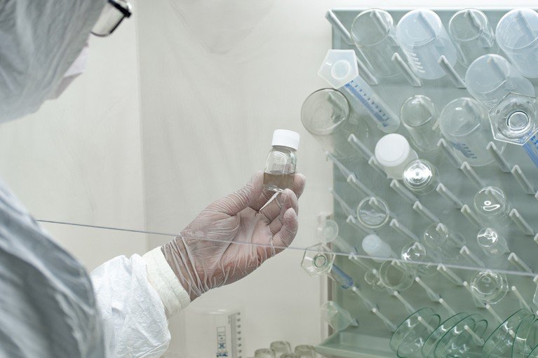 A researcher wearing PPE in a lab holds a small bottle of clear solution in front of a board of clean glass flasks.