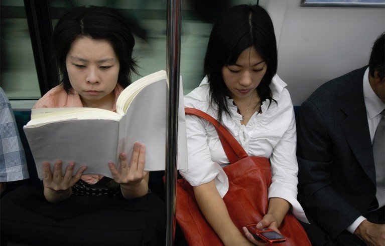 Seated train passengers during a commute, including a woman reading a book, another woman using a mobile phone, and a suited man. Vertical handrail divides carriage scene.