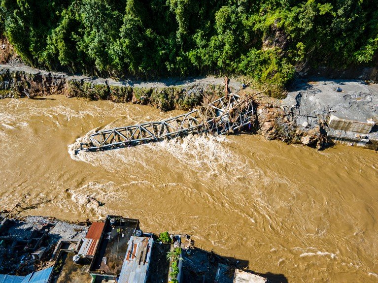 Aerial view of the aftermath of flooding along a river, with damage to homes and roads, and a collapsed bridge.
