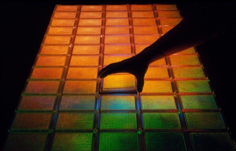 A hand interacting with a large rectangular grid of illuminated panels, likely an interactive light installation. Orange, yellow, green and blue squares form a graduated colour pattern against a black background.