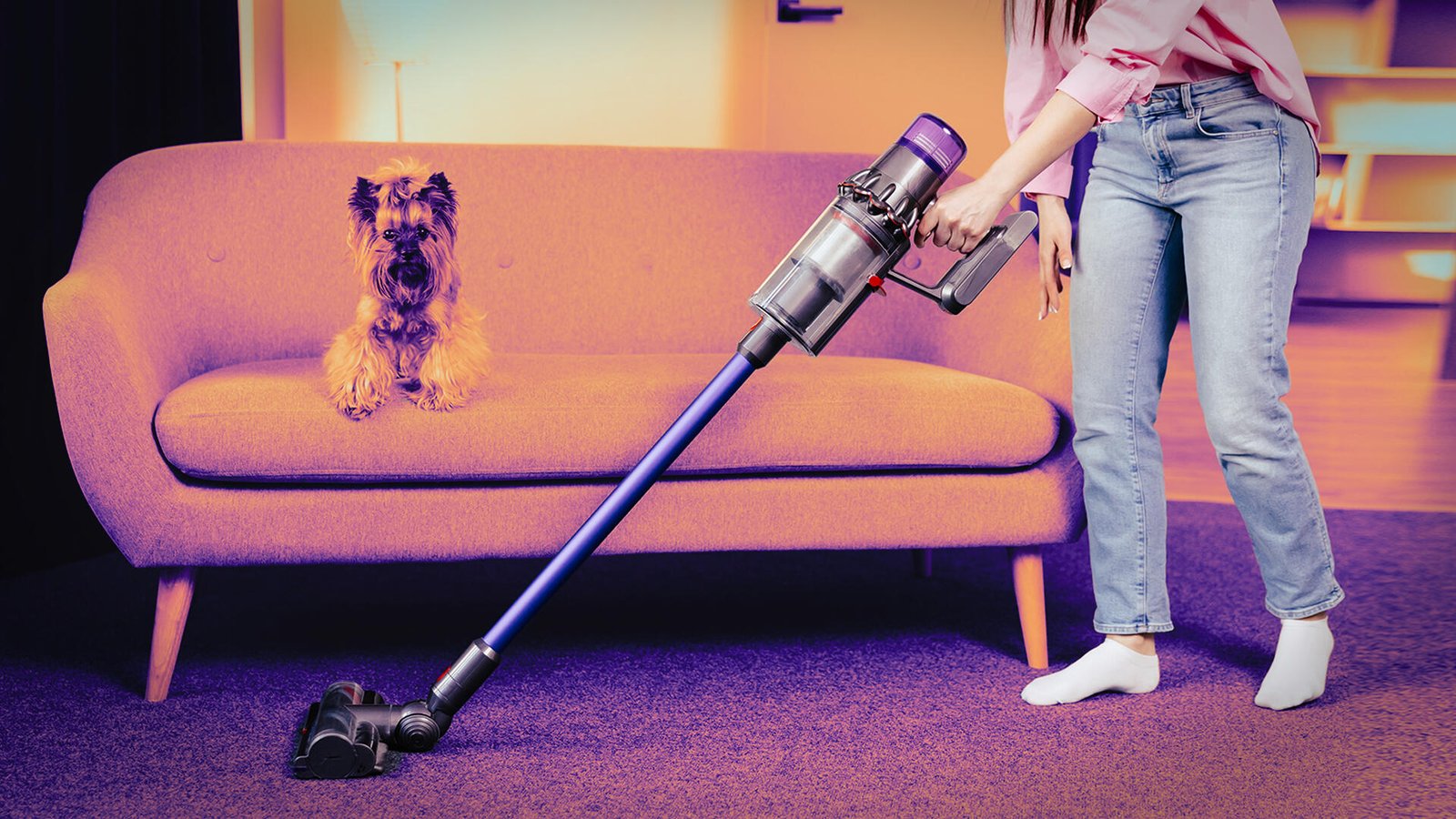 A woman pushes a cordless vacuum in front of a couch while a dog sits on the couch.