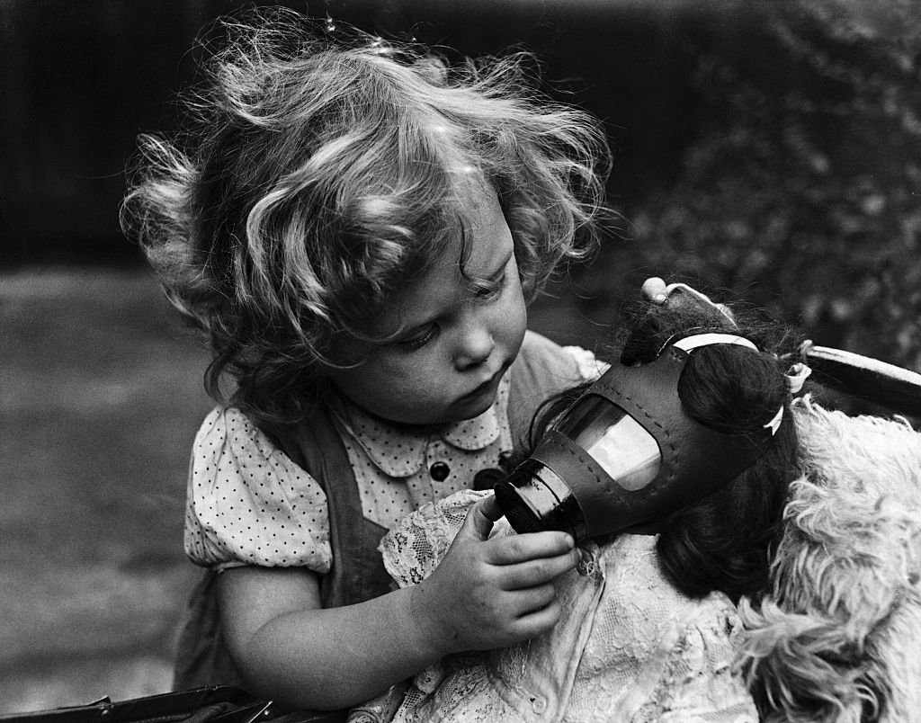 Little girl puts gas mask on doll in black and white vintage photograph.